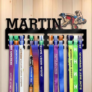 May include: Black metal medal display with the name "MARTIN" cut out. A wrestling figure is above the name. Colorful ribbons hang below, displaying medals. The display is mounted on a light wood-grain background.