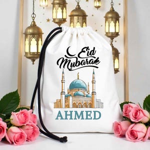 May include: White drawstring bag with a mosque illustration, blue domes and minarets. The bag has the text "Eid Mubarak" and the name "AHMED" in teal. Black drawstring closure. Pink roses and hanging lanterns in the background.