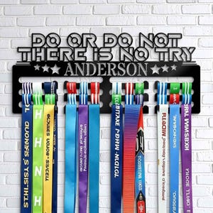 May include: A black metal medal display rack with the text "Do or do not, there is no try" and "Anderson" in white. The rack has 10 hooks for hanging medals. The rack is mounted on a white brick wall.