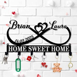 May include: Black metal key holder with the names "Brian" and "Laura" above the phrase "HOME SWEET HOME". The design includes an infinity symbol with a heart, the date 14.02.2025, and hooks for keys. The background is a white brick wall.