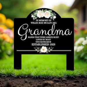 May include: Black metal garden sign with white flowers and the text "In memory of Willie Mae Mullin aka Grandma Hands that were always busy! Loved by many Our Matriarch Established 1929"