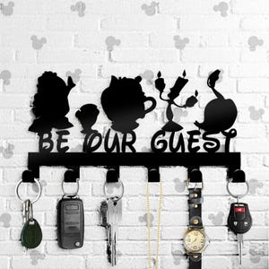 May include: Black metal key holder with silhouettes of characters from Beauty and the Beast. The holder has six hooks and the text "Be Our Guest".