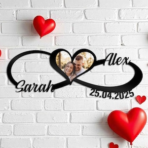 Puede incluir: Signo de infinito de metal negro con dos marcos de fotos en forma de corazón, los nombres "Sarah" y "Alex" y la fecha "25.04.2025". Globos rojos en forma de corazón en el fondo. El letrero está montado en una pared de ladrillos blancos.