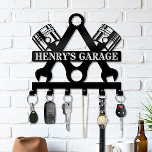 May include: Black metal key holder with the text "HENRY'S GARAGE". The design includes crossed pistons, wrenches, and key hooks. Various keys, a watch, and a necklace hang from the hooks. The background is a white brick wall.