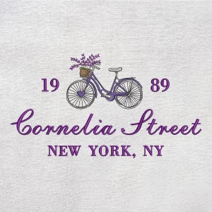 May include: Embroidered design featuring a purple bicycle with a basket of lavender flowers. The text reads "Cornelia Street, New York, NY" with the numbers "19" and "89" on either side of the bicycle. The design is on a white textured background.