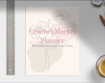 Op de afbeelding: Een crèmekleurige Crochet Market Planner met een bloemenlijnkunstontwerp en de tekst "Handmade Inventory & Profit Tracker". Een metalen liniaal staat links. Een kopje koffie en een pen staan rechts.