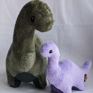 May include: Two plush dinosaur toys. One is a large, olive green Brontosaurus with a long neck and small black eyes. The other is a smaller, lavender dinosaur with a similar design. Both are made of soft, fuzzy material and are suitable for children.
