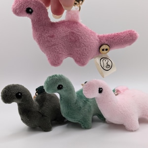 May include: Plush dinosaur keyrings in shades of pink, green, and grey. A pink dinosaur is held up by a gold-coloured clasp. Each dinosaur has a button and a small tag attached. The keyrings are soft and cuddly.