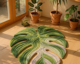 May include: A large, leaf-shaped rug in shades of green and white, resembling a Monstera plant leaf. The rug is placed on a wooden floor, with potted plants and natural light in the background. The rug's dimensions are approximately 70 inches long and 60 inches wide.