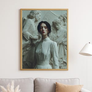 May include: A framed art print featuring a woman in a white dress, standing before a classical relief sculpture. The artwork is set in a gold frame and hangs above a gray sofa with a brown pillow. A white vase with pampas grass sits on a table.