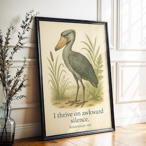 Op de afbeelding: Een ingelijste print met een gedetailleerde illustratie van een schoenbekooievaar in een moerasomgeving. Het kunstwerk is uitgevoerd in gedempte groen- en beigetinten, met de tekst "I thrive on awkward silence. Balaeniceps rex" onderaan.