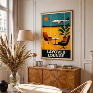May include: A framed art print featuring a retro travel-themed design. The artwork depicts an airplane flying past a window with a lounge scene, including chairs, a table, and the words "LAYOVER LOUNGE". The color palette includes orange, yellow, and teal.