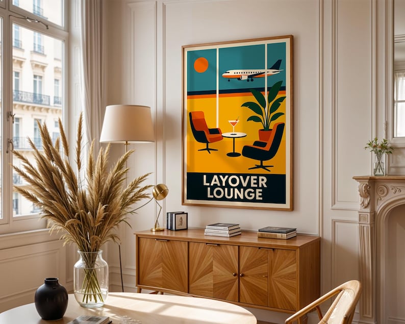 May include: A framed art print featuring a retro travel-themed design. The artwork depicts an airplane flying past a window with a lounge scene, including chairs, a table, and the words "LAYOVER LOUNGE". The color palette includes orange, yellow, and teal.