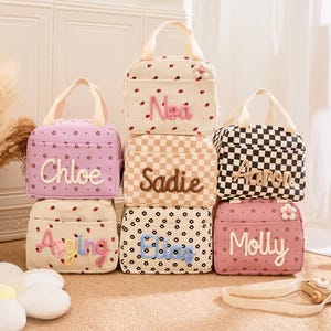 May include: A collection of personalized lunch bags in various colors and patterns. Each bag features a different name embroidered in a playful font. Designs include polka dots, checkerboard, and floral prints, with a small flower accent.