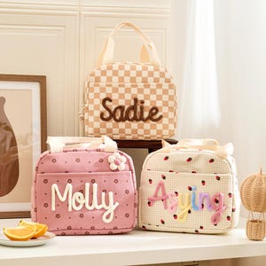 May include: Three personalized lunch bags in a soft color palette. One is checkered with "Sadie" embroidered, another is pink with "Molly", and the last is cream with "Aying". Each bag has a top handle.