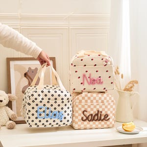 May include: Three personalized lunch bags. One bag is cream with black floral print and the name "Eliza" in blue. Another is cream with red ladybug print and the name "Noa" in pink. The third is tan checkered with the name "Sadie" in brown.