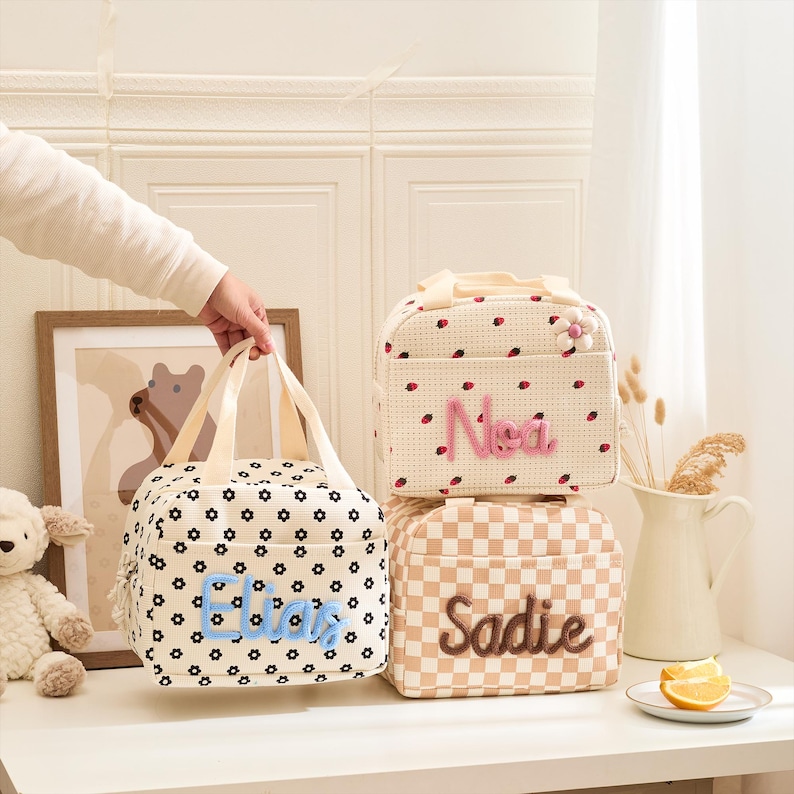 May include: Three personalized lunch bags. One bag is cream with black floral print and the name "Eliza" in blue. Another is cream with red ladybug print and the name "Noa" in pink. The third is tan checkered with the name "Sadie" in brown.