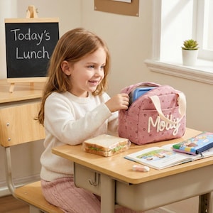 May include: A young girl smiles while opening a pink lunch bag with the name "Molly" on it. The bag is on a wooden desk with a sandwich, a book, and a pencil case. A small chalkboard reads "Today's Lunch".