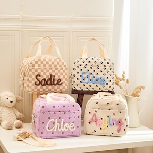 May include: Four personalized lunch bags with handles. One bag is tan and white checkered with "Sadie" embroidered. Another is white with black flower patterns and "Eliza" embroidered. The third is lavender with "Chloe" embroidered. The last is white with strawberries and "Aying" embroidered.