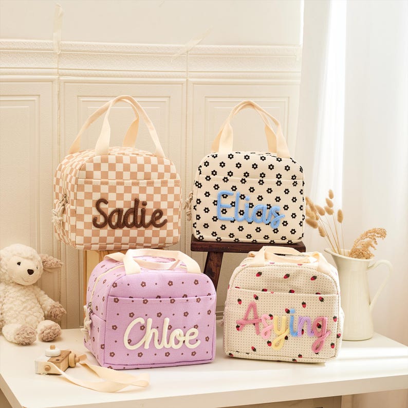 May include: Four personalized lunch bags with handles. One bag is tan and white checkered with "Sadie" embroidered. Another is white with black flower patterns and "Eliza" embroidered. The third is lavender with "Chloe" embroidered. The last is white with strawberries and "Aying" embroidered.