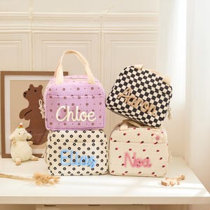 May include: Four personalized lunch bags stacked on a white surface. The bags feature different patterns: a lavender bag with the name "Chloe", a black and white checkered bag with "Aaron", a floral bag with "Eliza", and a strawberry-patterned bag with "Nia".