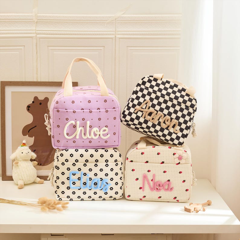 May include: Four personalized lunch bags stacked on a white surface. The bags feature different patterns: a lavender bag with the name "Chloe", a black and white checkered bag with "Aaron", a floral bag with "Eliza", and a strawberry-patterned bag with "Nia".