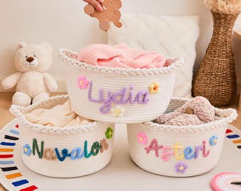 May include: Three white rope baskets with embroidered names and floral accents. The baskets are filled with soft items. The names are "Lydia", "Navalon", and "Asteri". A wooden toy is held above the baskets.