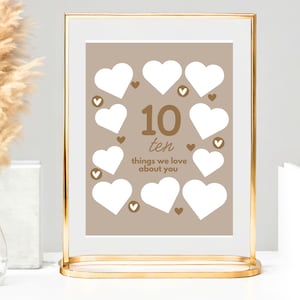 May include: A framed print in a gold metal stand. The print has a tan background with ten white heart shapes and brown heart accents. The text "10 ten things we love about you" is in brown. Pampas grass in a glass vase is on the left.