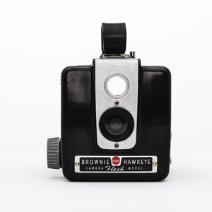 May include: A black Kodak Brownie Hawkeye Flash camera with a silver front panel and a red Kodak logo. The camera has a built-in flash and a viewfinder.