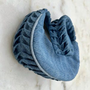 Op de afbeelding: Een blauwe denim hoofdband met een wit stiksel detail. De hoofdband is dubbelgevouwen en heeft een textuur, gerimpeld uiterlijk.