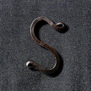 May include: A hand-forged, dark metal S-hook. The hook is made of a dark, possibly iron, metal with a textured surface. The S-shape has rounded ends, and the hook is set against a dark, textured background.