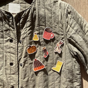 Op de afbeelding: Een groene gewatteerde jas met verschillende geborduurde patches, waaronder een pompoen, een peer, een laars, een hoed, een mok, een sok en een konijn.