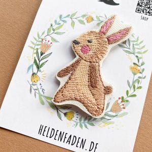 Peut inclure: Écusson brodé représentant un lapin de dessin animé avec des détails marron et rose. L'écusson est sur une carte blanche avec un motif de couronne florale et le texte "HELDENFADEN.DE" et "SHOP".