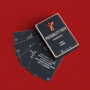 Puede incluir: Un mazo de cartas titulado "Possibility Path Decision Deck, Volume 1 Scenarios" con un logotipo de flecha hacia arriba. Las cartas son de color azul oscuro con texto blanco y un fondo rojo.
