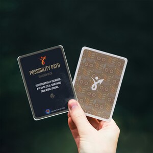 Puede incluir: Dos cartas del "Possibility Path Decision Deck" se sostienen en una mano. Una carta es azul oscuro con texto blanco y un logotipo de flecha naranja. La otra carta tiene un patr&oacute;n marr&oacute;n y beige.
