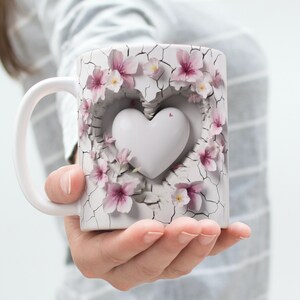 Könnte beinhalten: Weiße Keramiktasse mit einem rissigen Design und rosa Blumen, die ein weißes Herz umgeben.