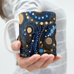Könnte beinhalten: Eine weiße Keramiktasse mit einem abstrakten Design in Blau und Gold. Das Design zeigt Kreise, Punkte und Wirbel.