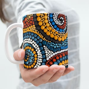 Könnte beinhalten: Weiße Keramiktasse mit einem Muster aus schwarzen, gelben, blauen, roten und weißen Punkten. Das Muster ähnelt der Aborigine-Kunst.