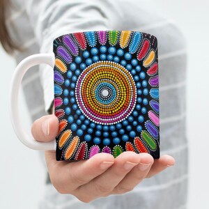 Könnte beinhalten: Eine weiße Keramiktasse mit einem bunten Punkt-Mandala-Design. Das Design zeigt einen zentralen Kreis mit konzentrischen Ringen aus Punkten in verschiedenen Farben, darunter Rot, Orange, Gelb, Grün, Blau, Lila und Pink. Der Hintergrund ist schwarz.