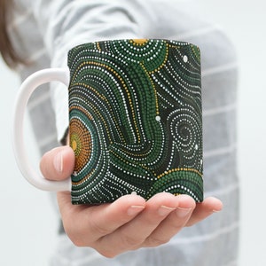 Könnte beinhalten: Eine weiße Keramiktasse mit einem abstrakten Muster in Grün, Gelb und Weiß. Die Tasse hat einen Henkel und wird von einer Hand gehalten.