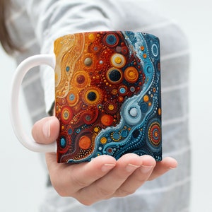 Puede incluir: Una taza de cerámica blanca con un diseño abstracto colorido. El diseño presenta una mezcla de colores naranja, amarillo, azul y marrón en un patrón de remolinos de puntos y círculos.