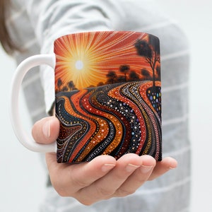 Puede incluir: Taza de cerámica blanca con un diseño abstracto colorido. El diseño presenta una puesta de sol con un sol, árboles y un paisaje de puntos naranja, rojo, negro y blanco.