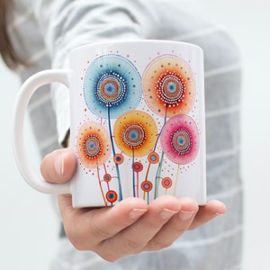 May include: White ceramic mug featuring a colourful dandelion design. The dandelions are painted in shades of blue, orange, yellow, and pink.