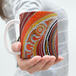 Könnte beinhalten: Eine weiße Keramiktasse mit einem farbenfrohen Design. Das Design zeigt ein Muster aus Punkten und Linien in Orange-, Rot-, Gelb- und Schwarztönen. Die Tasse wird in der Hand einer Person gehalten.
