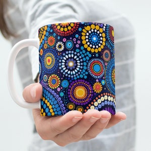 Könnte beinhalten: Eine weiße Keramiktasse mit einem farbenfrohen abstrakten Design mit Kreisen und Punkten in Blautönen, Gelb, Rot, Lila und Weiß auf dunkelblauem Hintergrund.