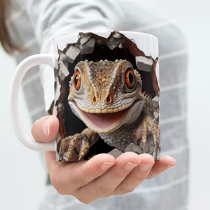 Puede incluir: Taza de cerámica blanca con un lagarto sonriente que rompe por el lado. El lagarto es marrón y tiene ojos naranjas. La taza tiene un asa en el lado izquierdo.