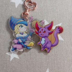 Może przedstawiać: Zawieszka z przezroczystego akrylu przedstawiająca kreskówkową dziewczynkę z blond włosami, ubraną w niebiesko-biały kapelusz czarownicy i różową sukienkę. Trzyma żółtą różdżkę. Zawieszka jest przymocowana do złotego breloka. Drewniana zawieszka przedstawiająca fioletowego kreskówkowego kota z czerwonymi oczami jest również przymocowana do breloka.