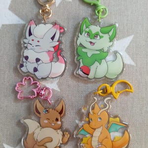 Puede incluir: Cuatro llaveros de personajes de dibujos animados coloridos con respaldo de plástico transparente. Los personajes son un zorro blanco, un gato verde, un Eevee marrón y un Charizard naranja.