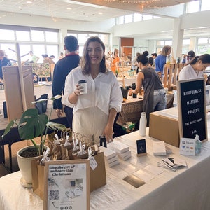 May include: A table display of self-care products with a sign that reads "Give the gift of self care this Christmas". The products include small brown paper bags, boxes of cards, and a sign that reads "Mindfulness Matters Project The Daily Cards 13.5".