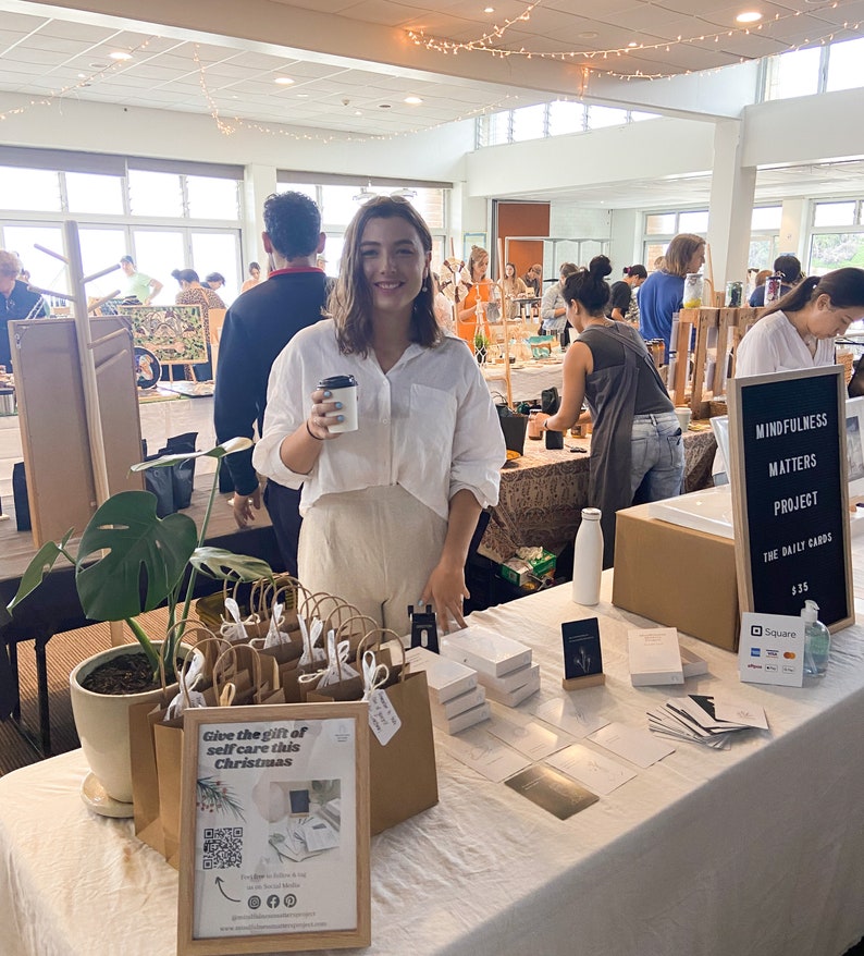 May include: A table display of self-care products with a sign that reads "Give the gift of self care this Christmas". The products include small brown paper bags, boxes of cards, and a sign that reads "Mindfulness Matters Project The Daily Cards 13.5".